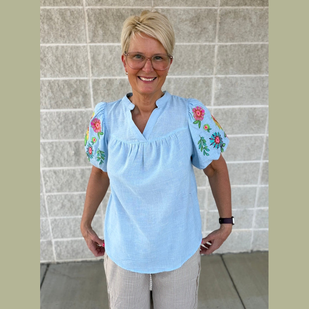 Woman with short blonde hair smiling, wearing a light blue top with embroidered puff sleeves and striped pants.