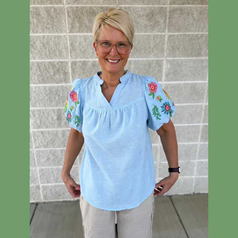 Woman wearing a light blue v-neck top with floral embroidered puff sleeves and striped pants against a brick wall.