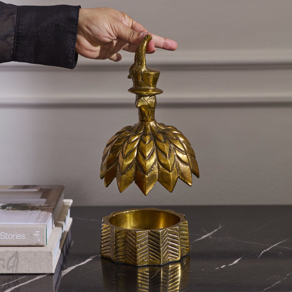 Person holding open the lid of an antique gold decorative box shaped like a bell featuring a feathered bird top hat handle.