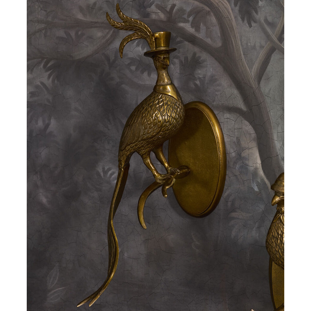 Vintage brass bird wall sconce wearing a top hat with feathers, mounted on a textured dark wall.