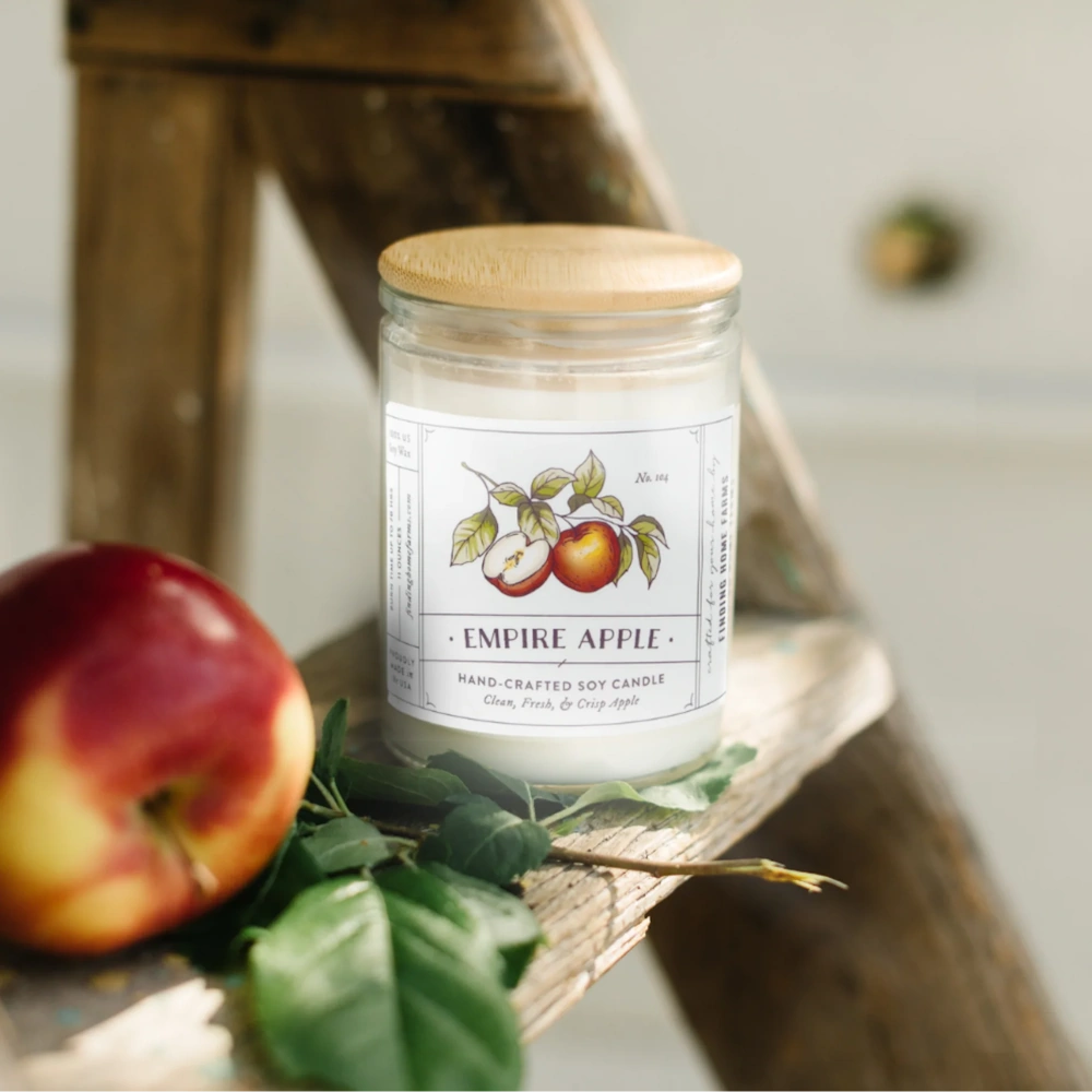Hand-crafted soy candle jar with wooden lid next to a fresh red apple resting on green leaves.