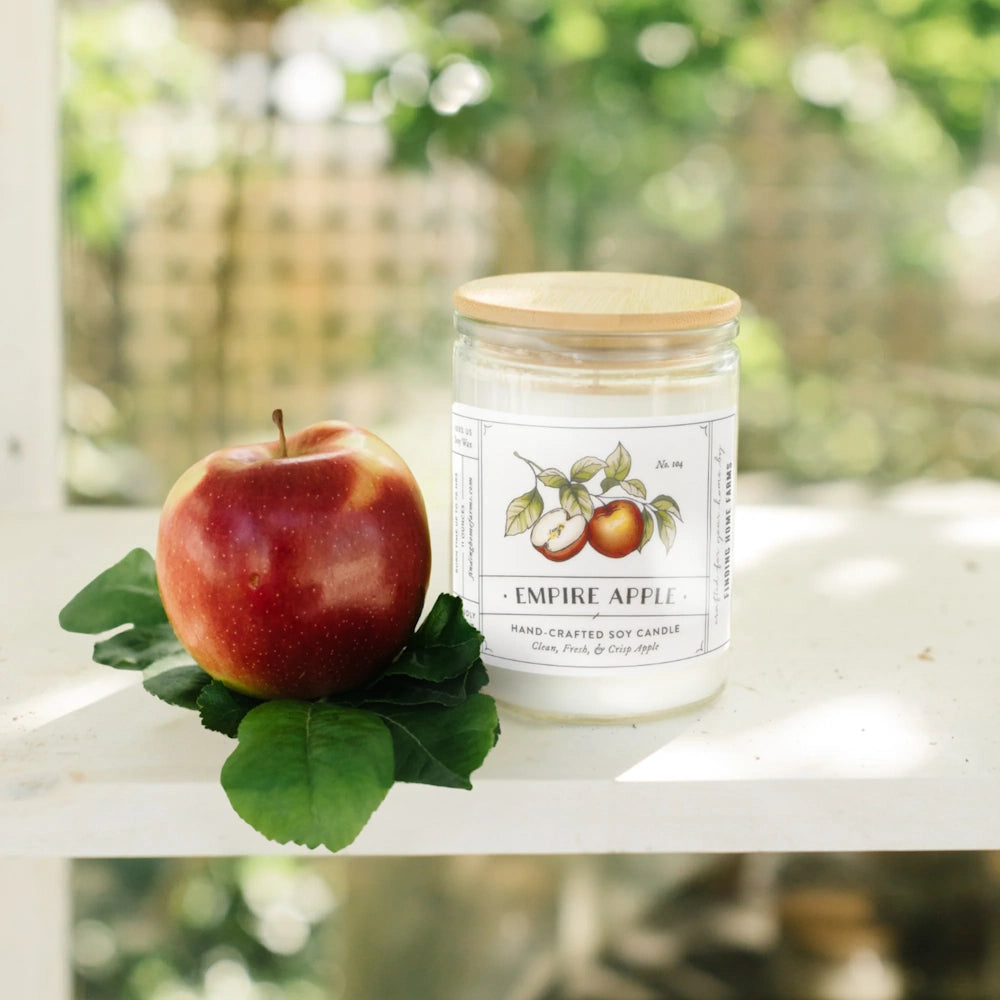 Empire Apple soy candle with a wooden lid on a rustic wooden ladder next to a fresh red apple.