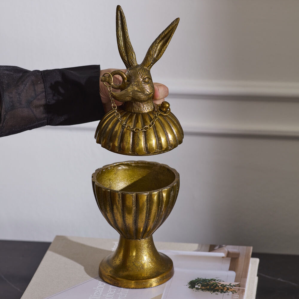 Person holding open the lid of an antique gold decorative box shaped like a pedestal urn with a mustached rabbit handle.