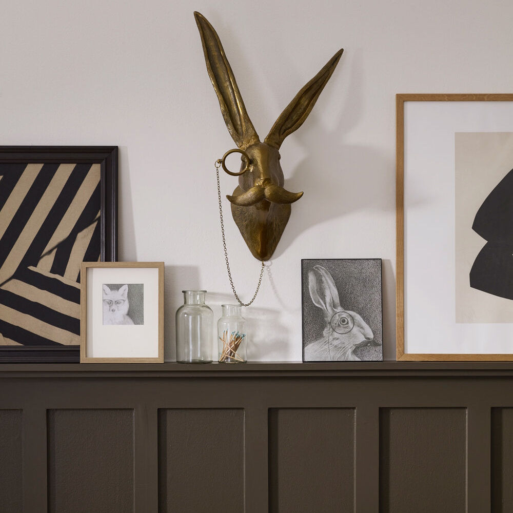 Brass hare head wall mount with a monocle and mustache, flanked by framed art on a ledge.