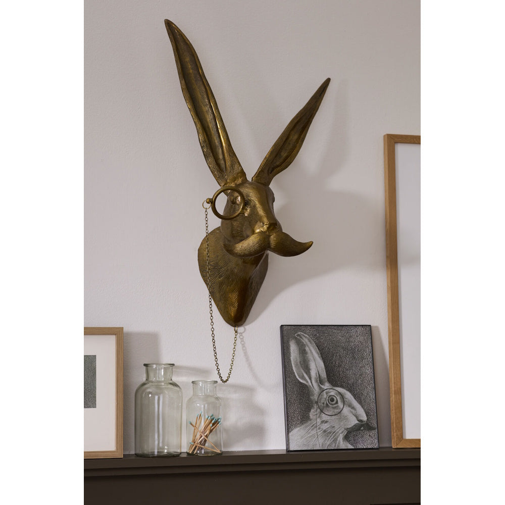 A gold-toned hare bust sculpture with long ears, a monocle, and a mustache mounted on a wall above a shelf.