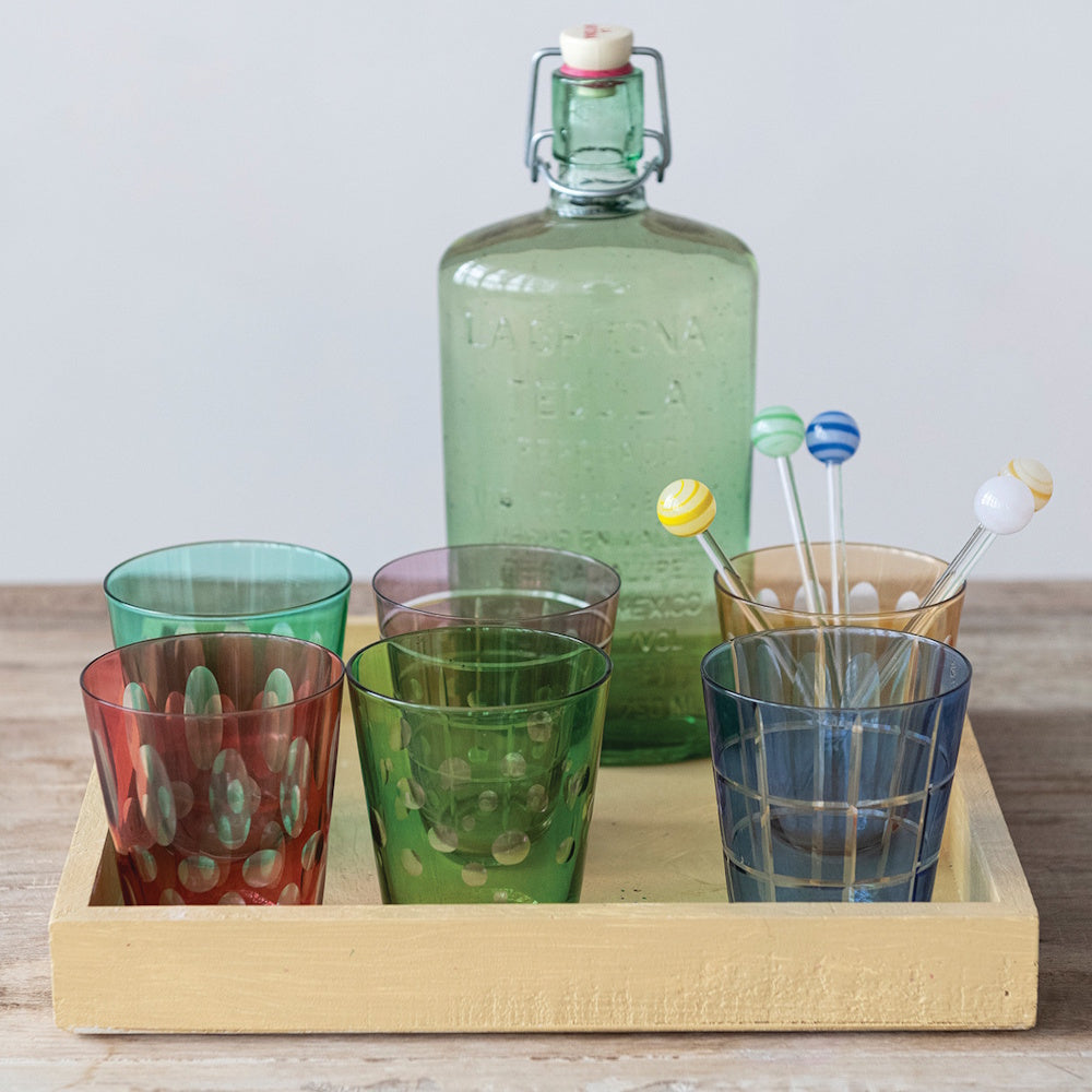 Colorful etched drinking glasses and glass swizzle sticks on a tray with a vintage bottle.