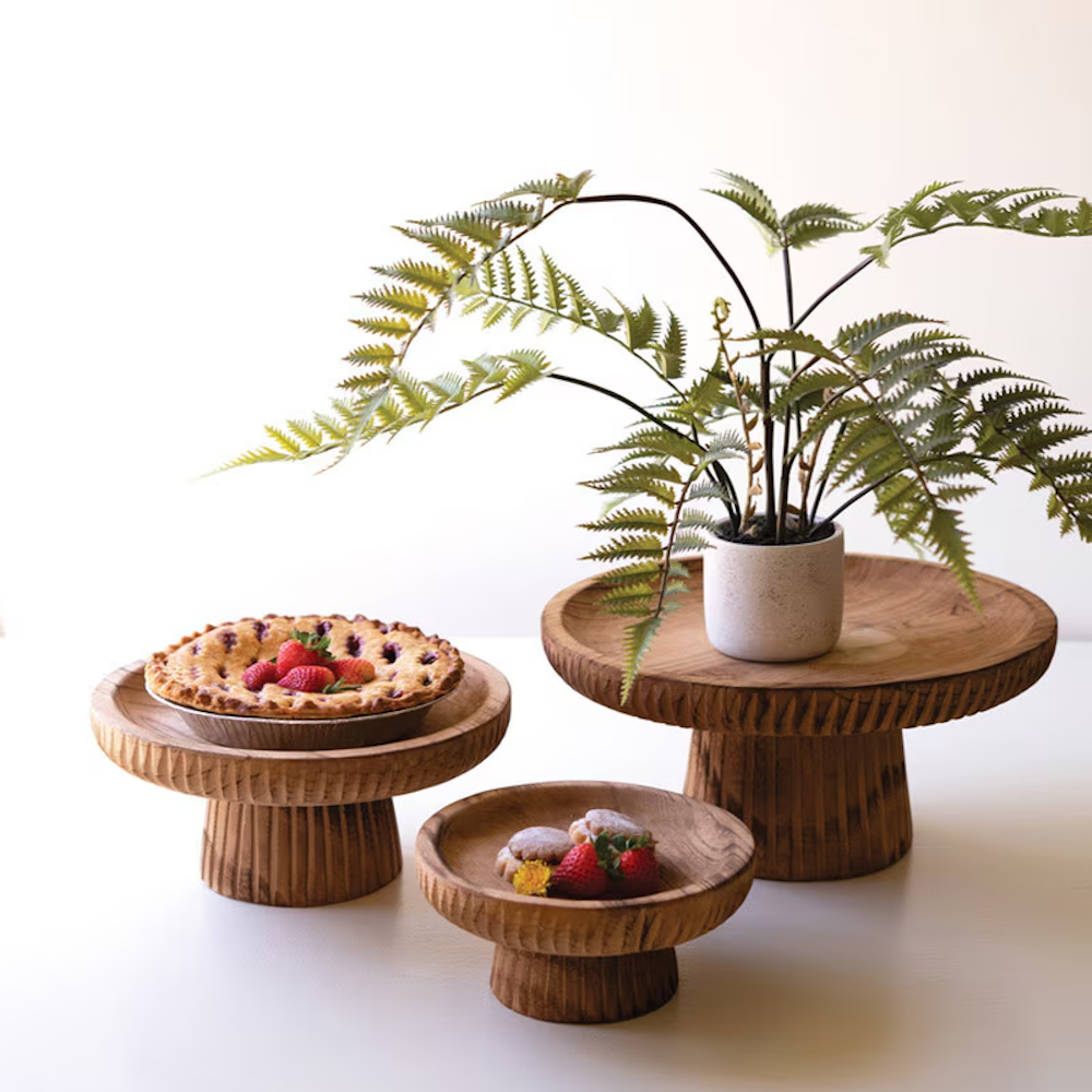 Trio of carved wooden pedestal stands holding pastries, a pie, and a potted fern against white background.