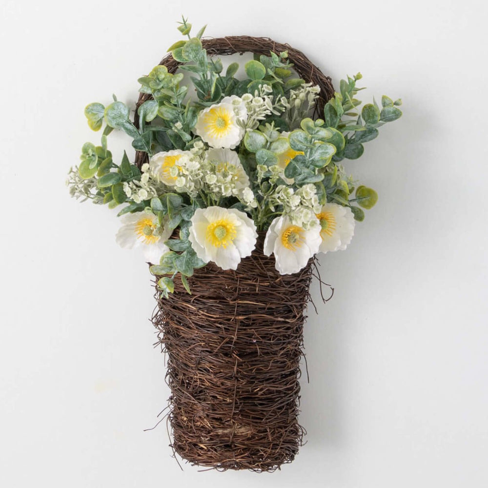 Woven twig hanging basket filled with artificial white poppies and green eucalyptus stems against a white wall.