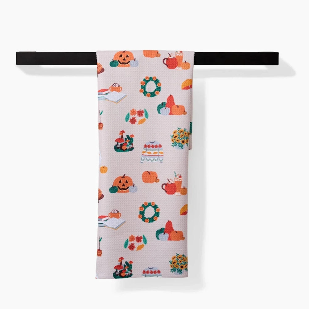 Light beige tea towel with a colorful pattern of fall icons hanging over a black bar.