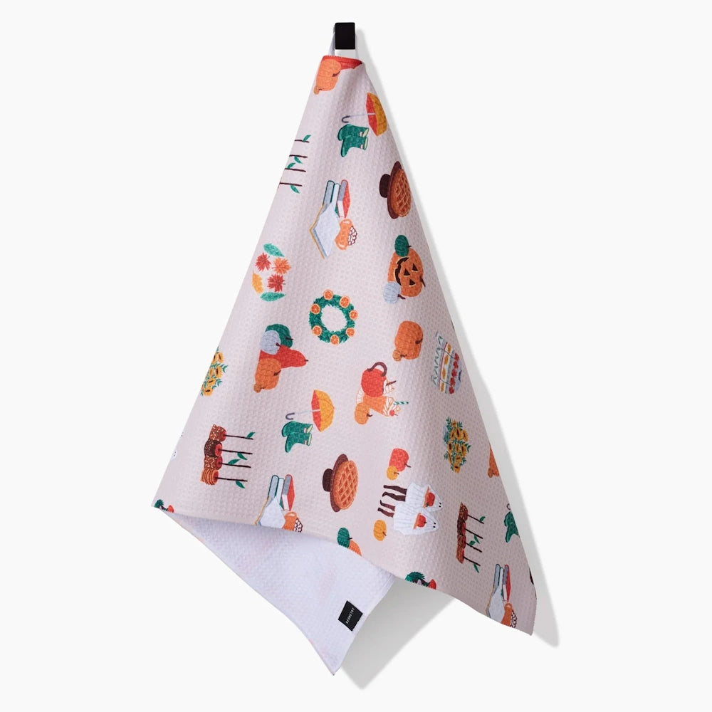 A hanging waffle-weave kitchen towel featuring a pattern of autumn items like pumpkins, pies, books, and boots.
