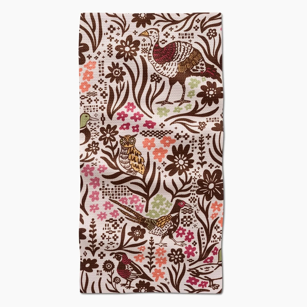 A white kitchen towel laid flat, featuring a folk-art pattern of autumn birds, flowers, and foliage.