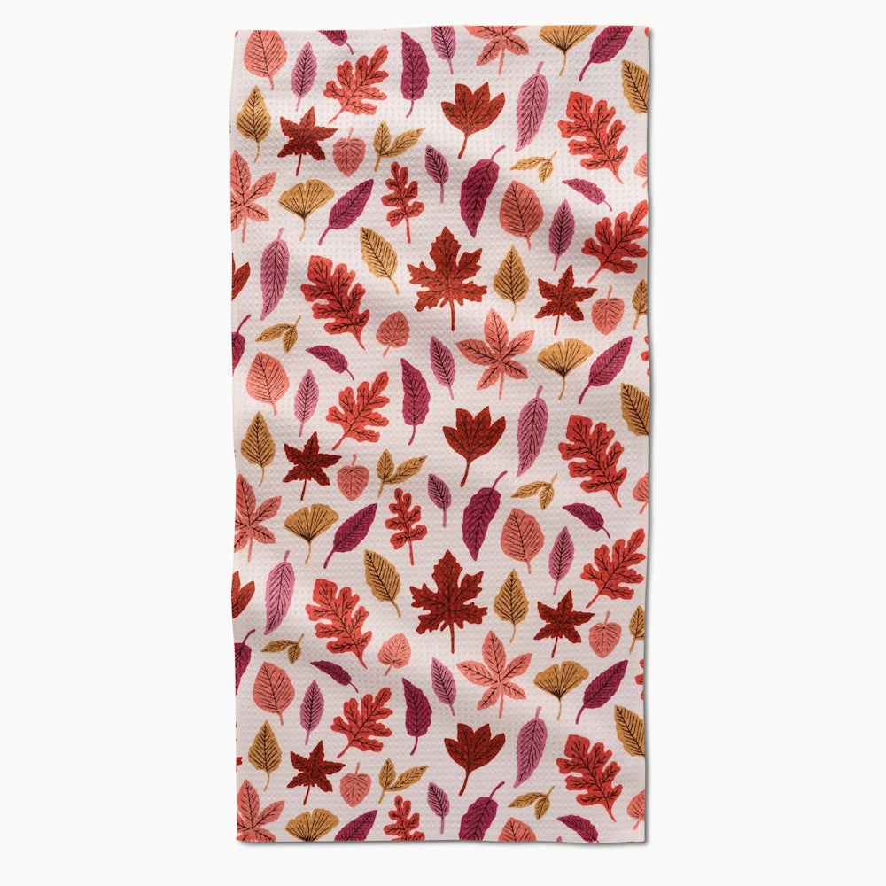 A white waffle-weave kitchen towel featuring a colorful pattern of scattered autumn leaves in red, orange, and purple.