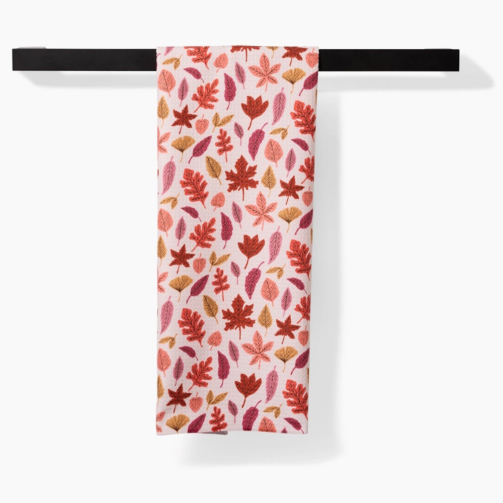 Pink kitchen towel with a repeated pattern of autumn leaves hanging from a modern black bar.