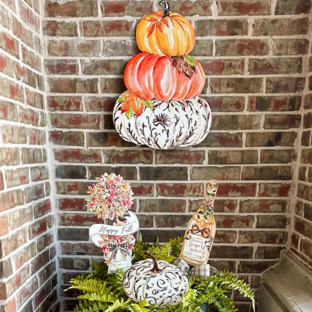 Autumn porch display with a stacked pumpkin sign on a brick wall above real pumpkins and ferns.