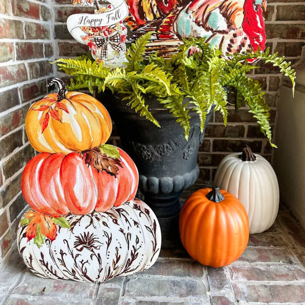 A stacked pumpkin sign next to real pumpkins and a potted fern as part of a festive autumn porch display.