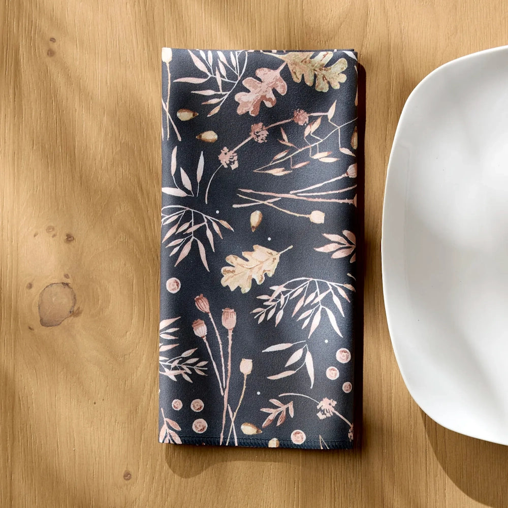 Folded dark gray dinner napkin with a botanical pattern of autumn leaves and stems next to a white plate.