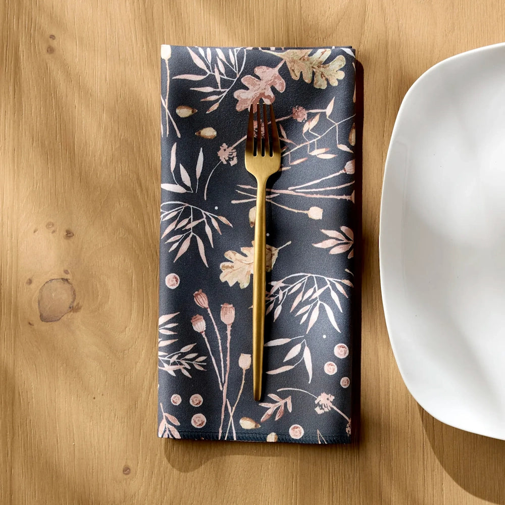 A dark patterned cloth napkin featuring an autumn botanical design with a gold fork on top next to a white plate.