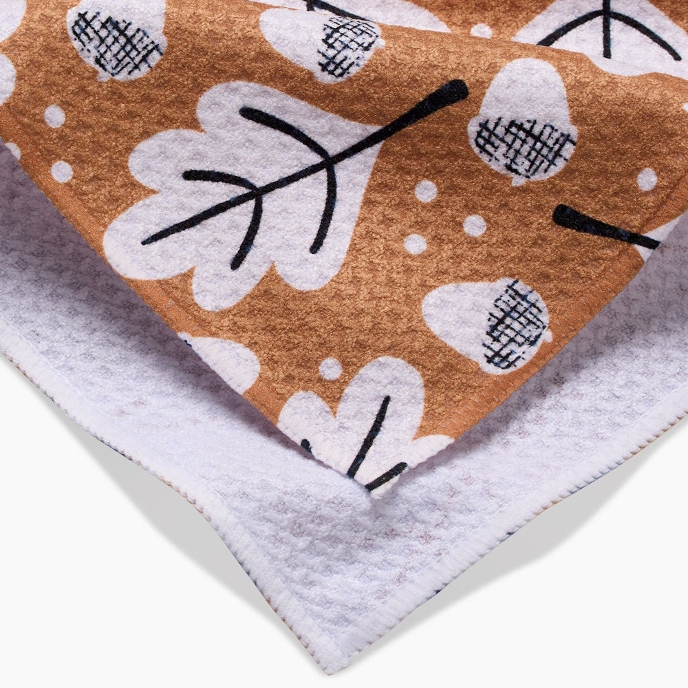 Close-up of a brown tea towel featuring a white and black pattern of falling oak leaves and acorns.