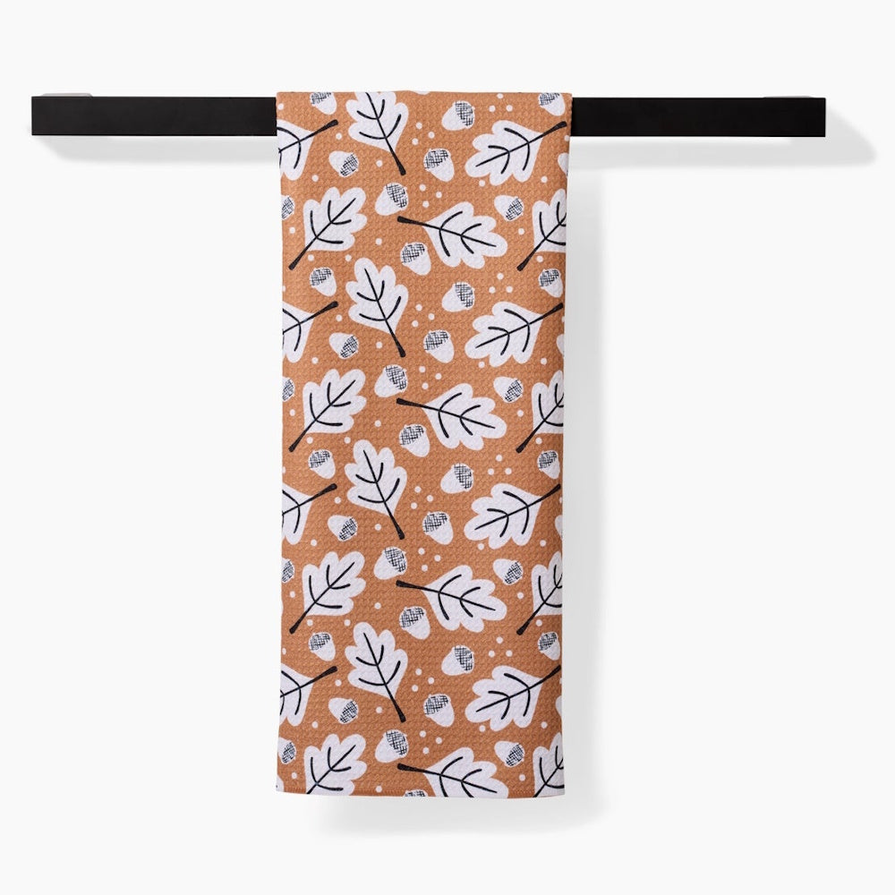 A tan kitchen towel patterned with white oak leaves and acorns hangs from a black modern towel bar.