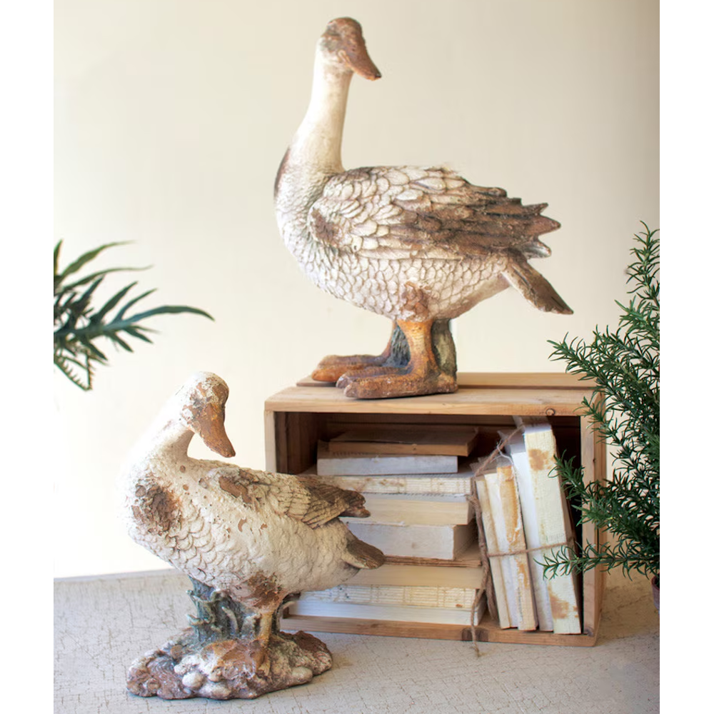 Two rustic duck statues with a distressed finish displayed near a wooden crate holding books.
