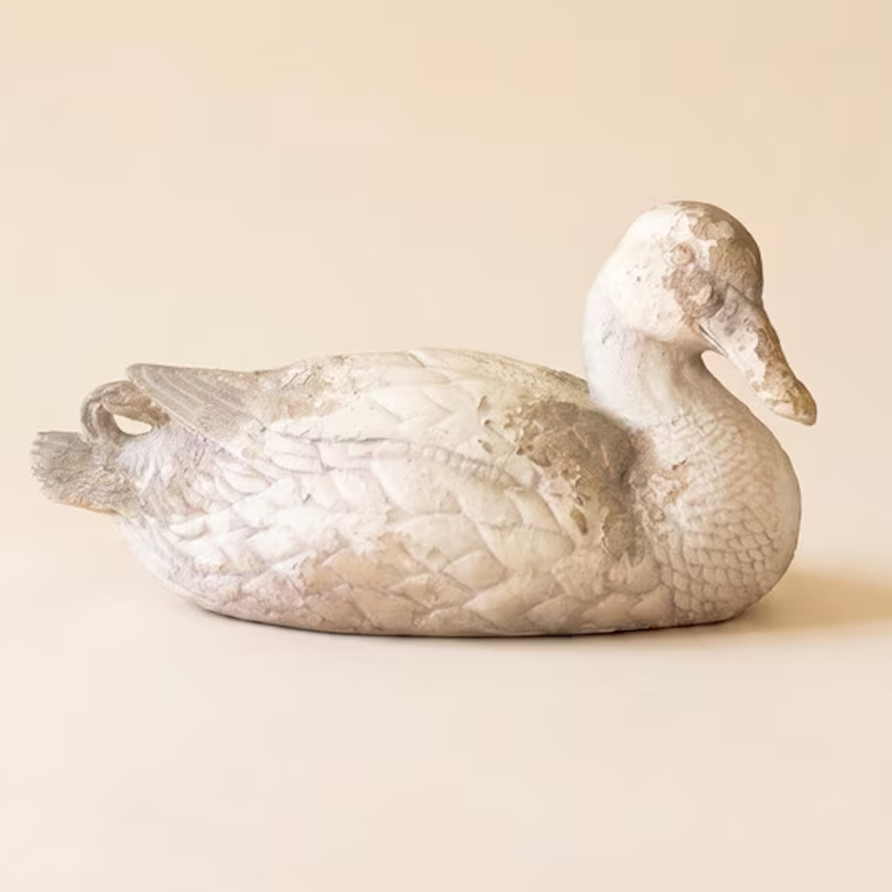 Weathered white pintail duck statue resting on a plain beige studio background.