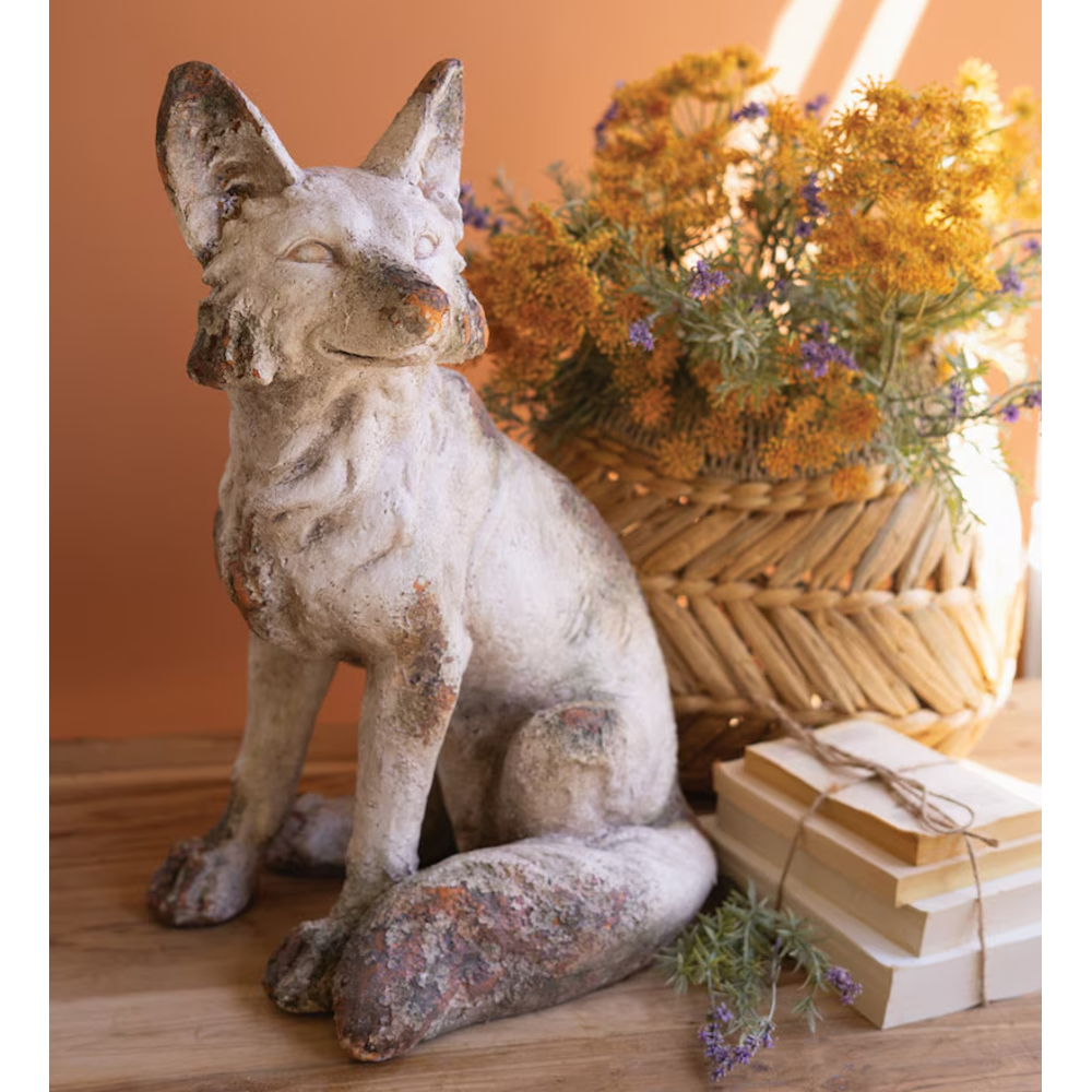 Weathered fox statue sitting next to a stack of books and a woven basket of dried flowers.