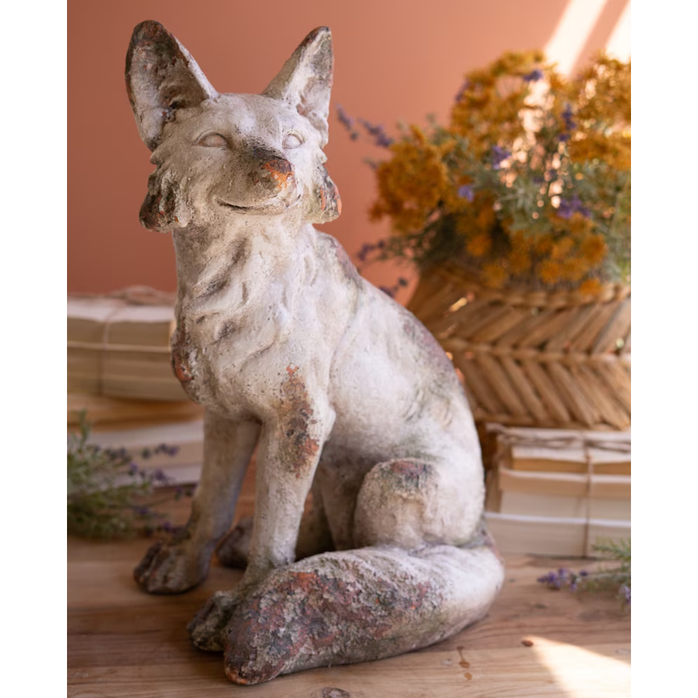 Weathered, faux-concrete sitting fox statue displayed on a wooden surface with stacks of books.