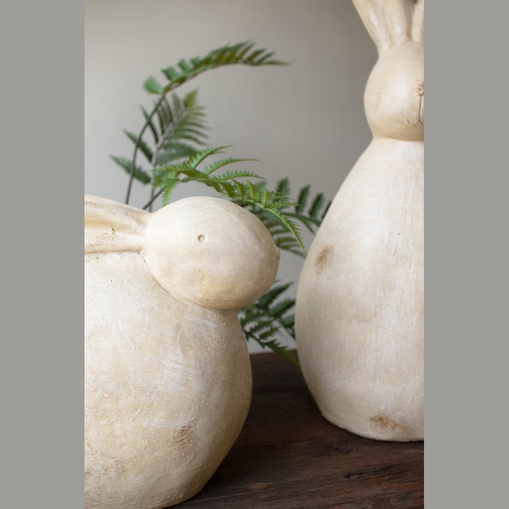 Two faux stone rabbit sculptures with a weathered cream finish and green fern accents on a rustic wooden surface.