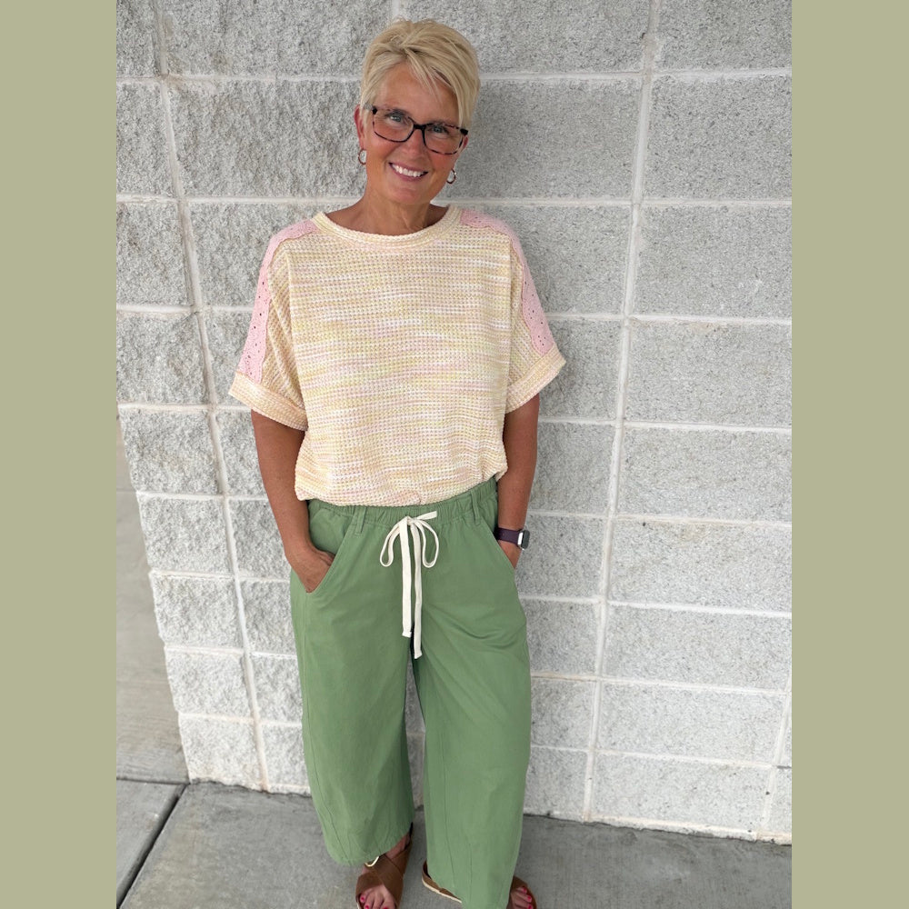 A woman smiling in front of a brick wall wearing a yellow textured top and green wide-leg drawstring pants.