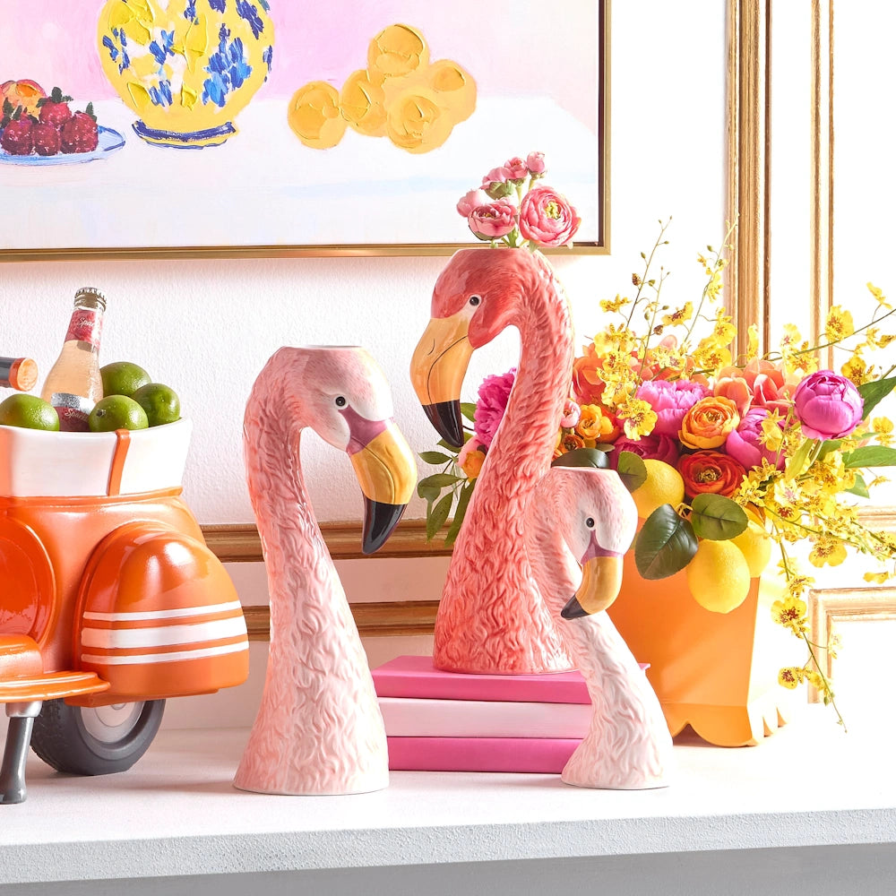 Three flamingo-shaped vases filled with colorful flowers sit on a shelf. Nearby, an orange scooter holds limes, against a bright, playful painting.