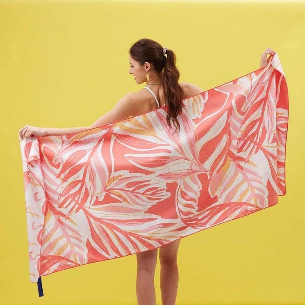 A woman wraps a pink and orange tropical leaf print beach towel around her shoulders against a yellow background.