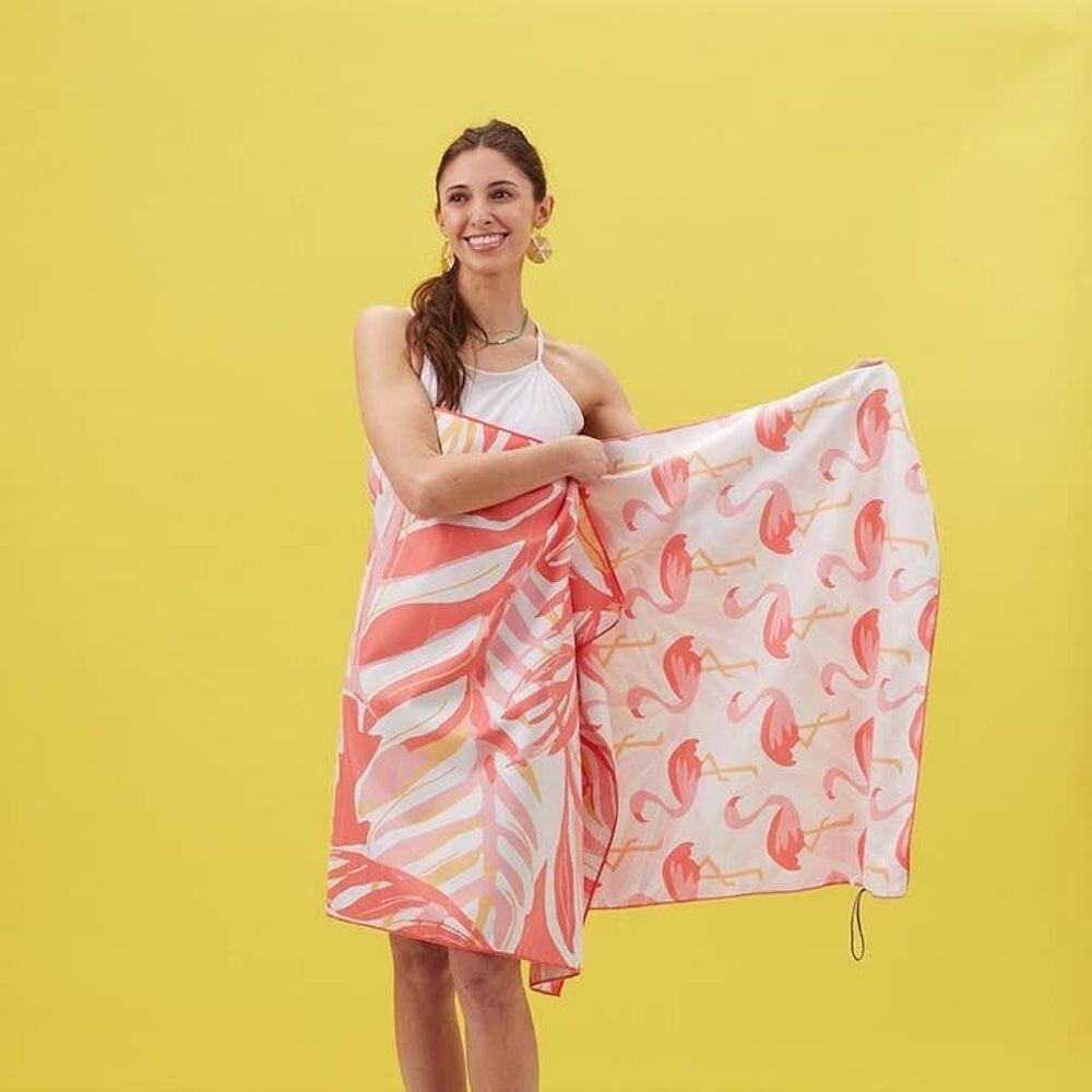 A smiling woman wraps herself in a beach towel featuring a reversible flamingo and leaf print pattern.