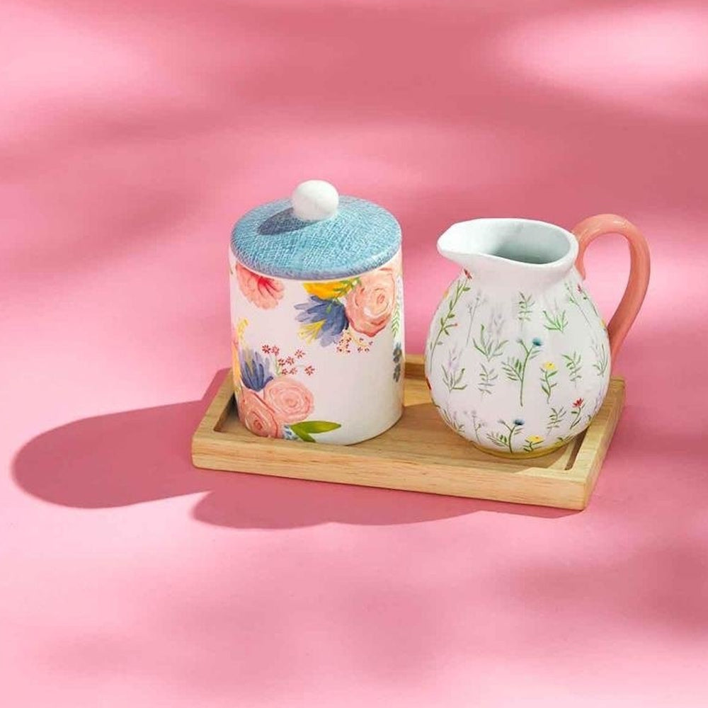 Floral ceramic cream and sugar set on a small wooden tray, set against a vibrant pink background.
