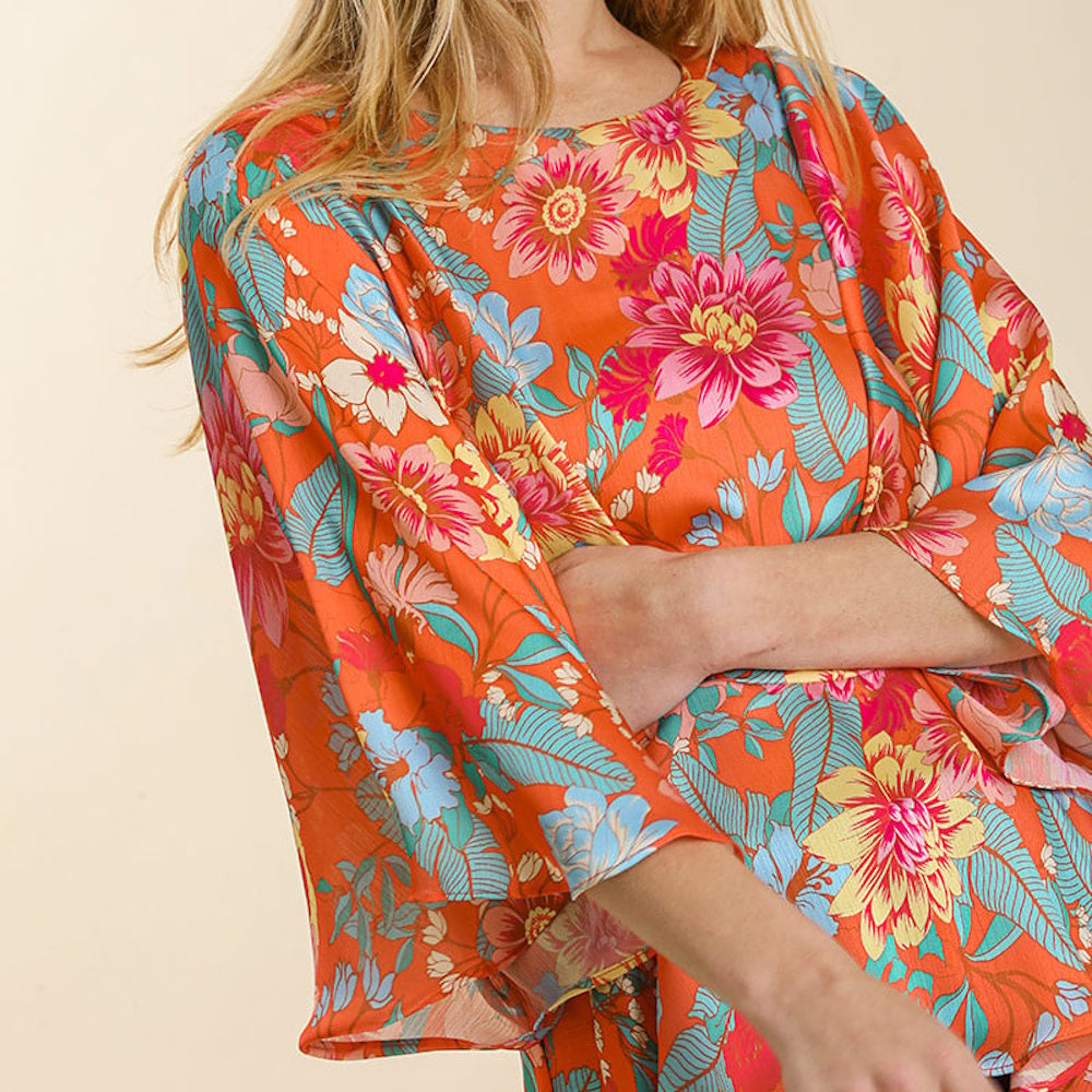 Woman wearing a bright orange top with a vibrant floral print and wide kimono-style sleeves.