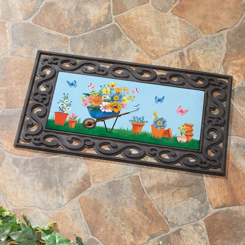 Decorative door mat featuring a floral wheelbarrow, potted plants, and butterflies on a blue background with a scroll border.
