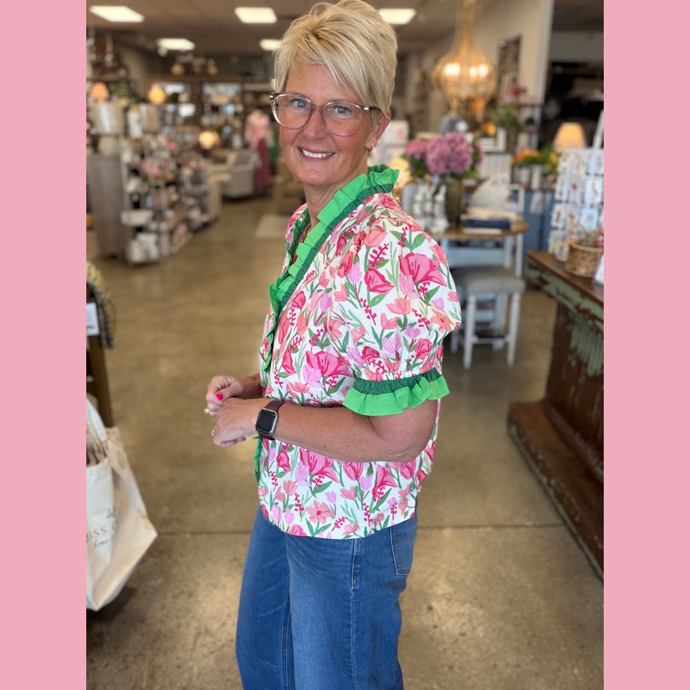 Woman in pink floral puff sleeve blouse with green ruffle trim and jeans standing in a retail boutique.