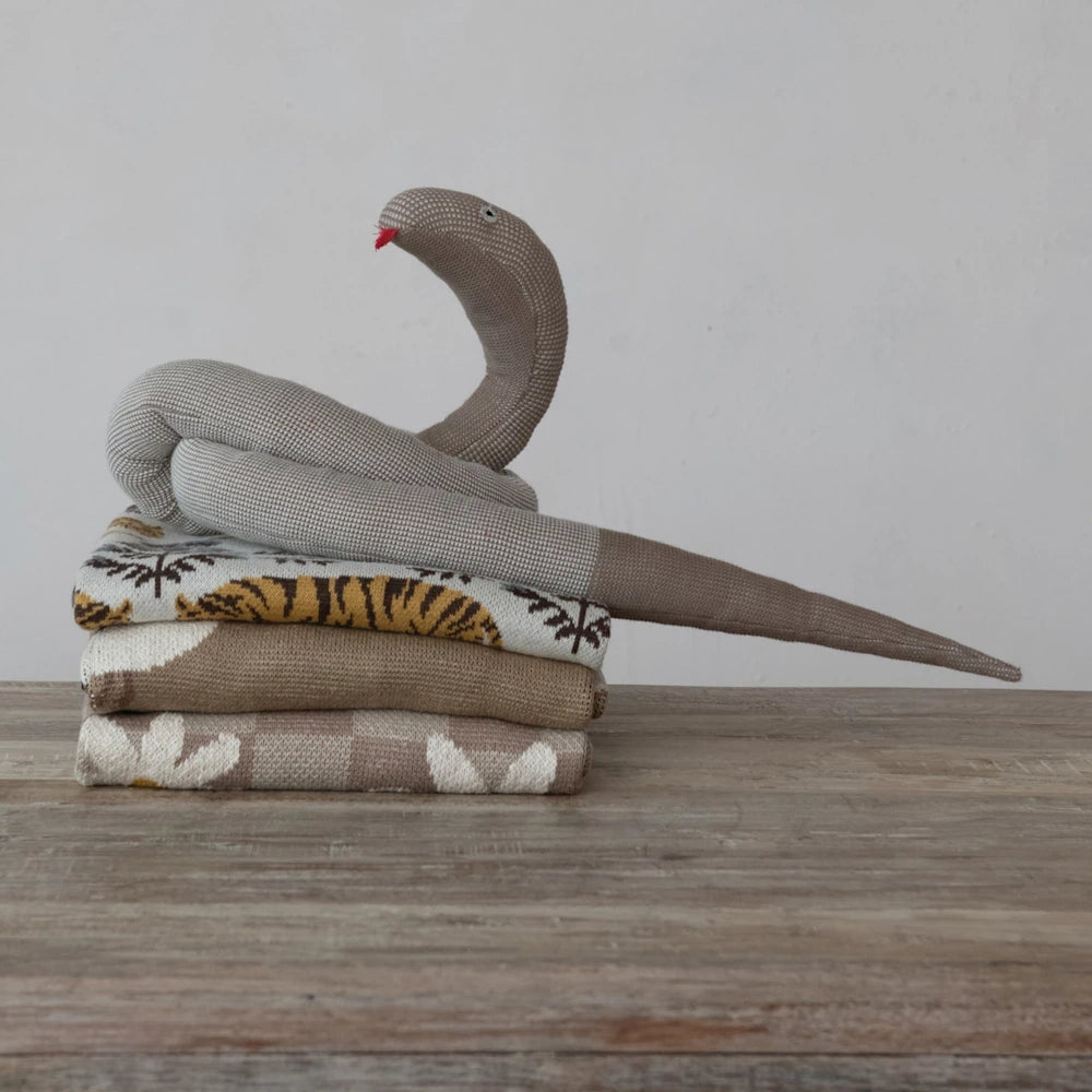 Knitted snake plush toy resting on a stack of patterned cotton baby blankets on a wooden table.