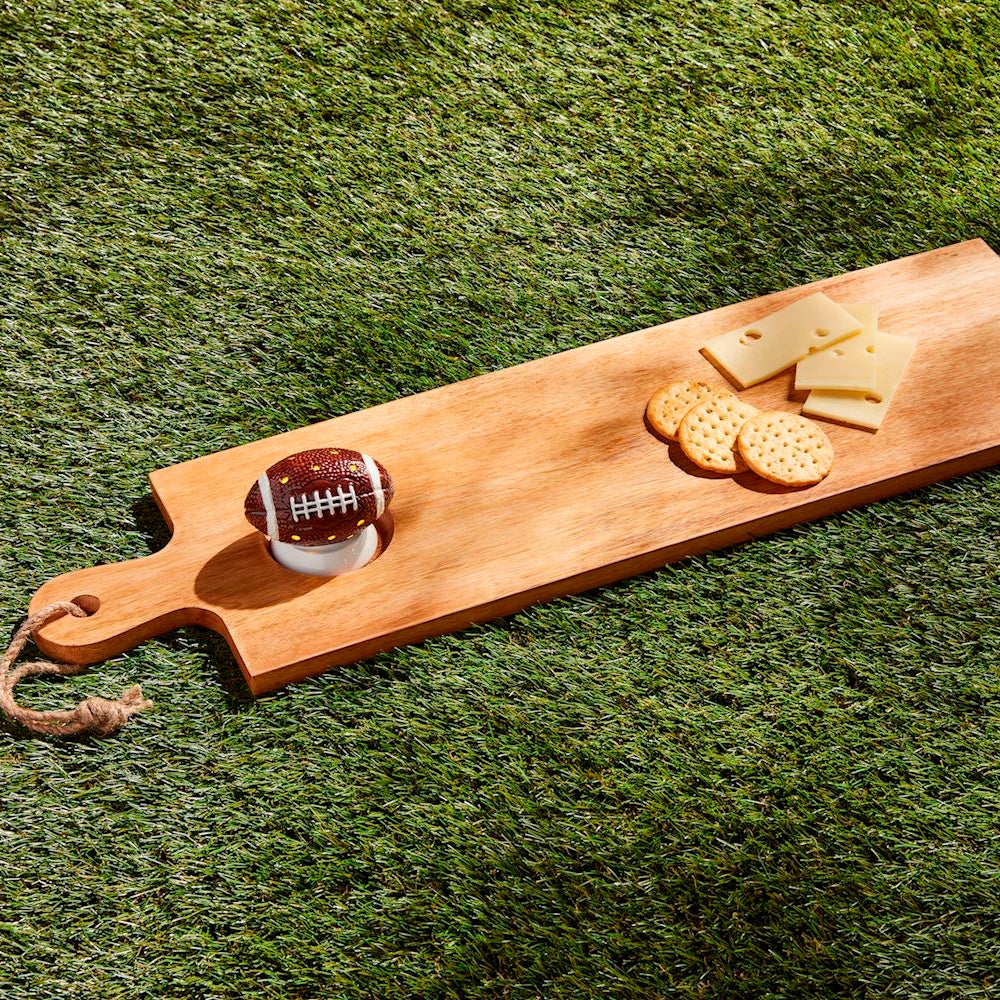 Wooden serving board on artificial turf featuring a mini football accent, cheese slices, and crackers.