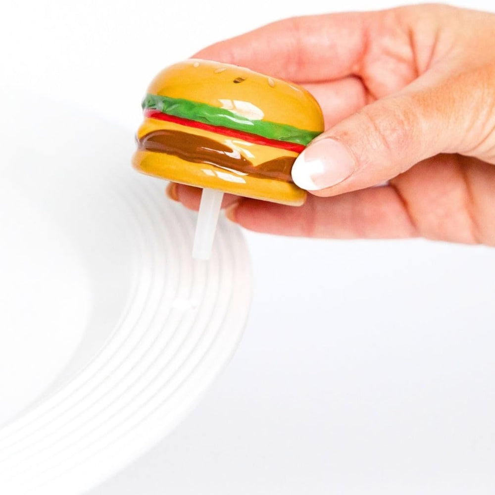 A hand holding a burger-themed Nora Fleming decorative ceramic mini over a white rimmed serving platter.