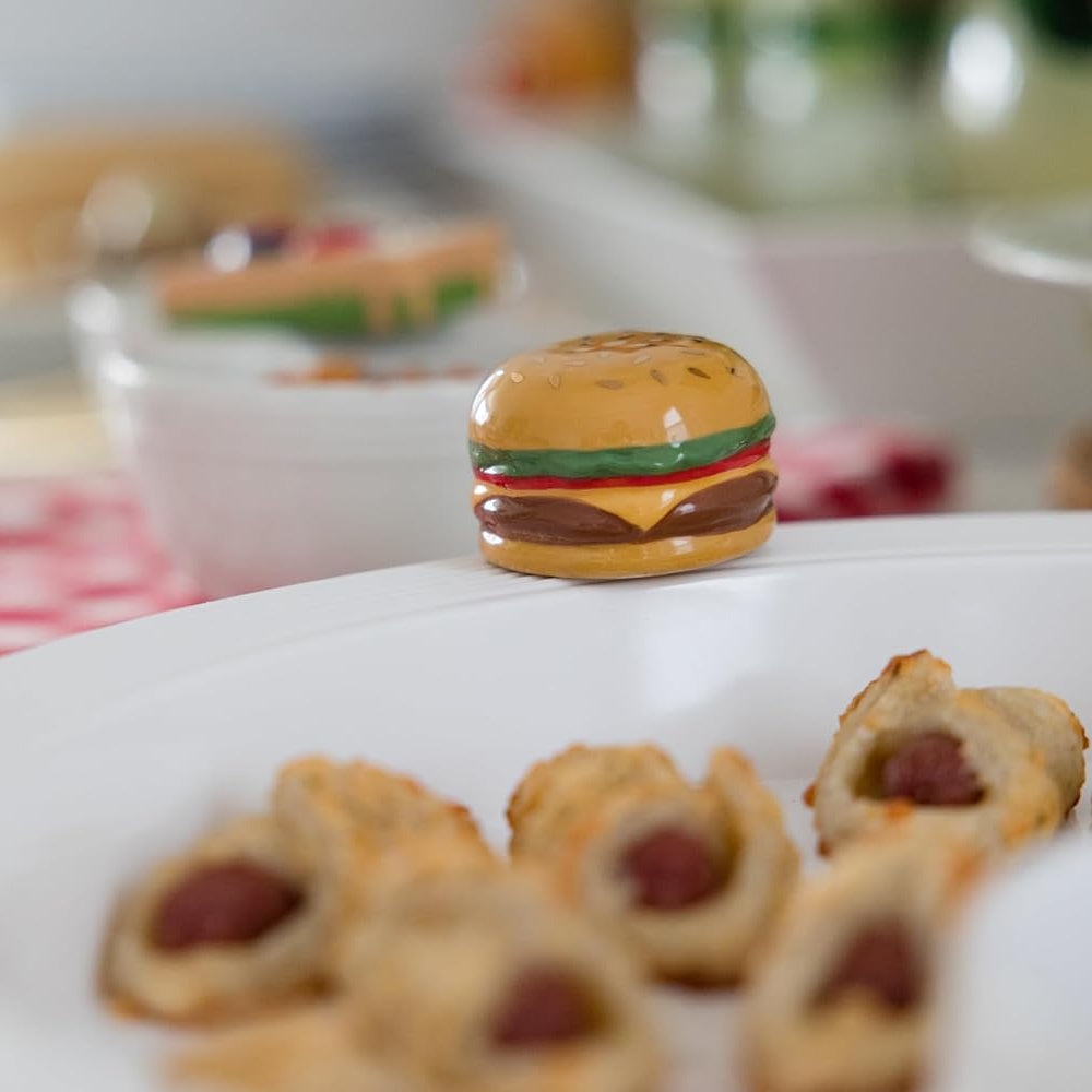 Close-up of a decorative ceramic burger mini charm attached to the rim of a white serving platter with pigs in a blanket.