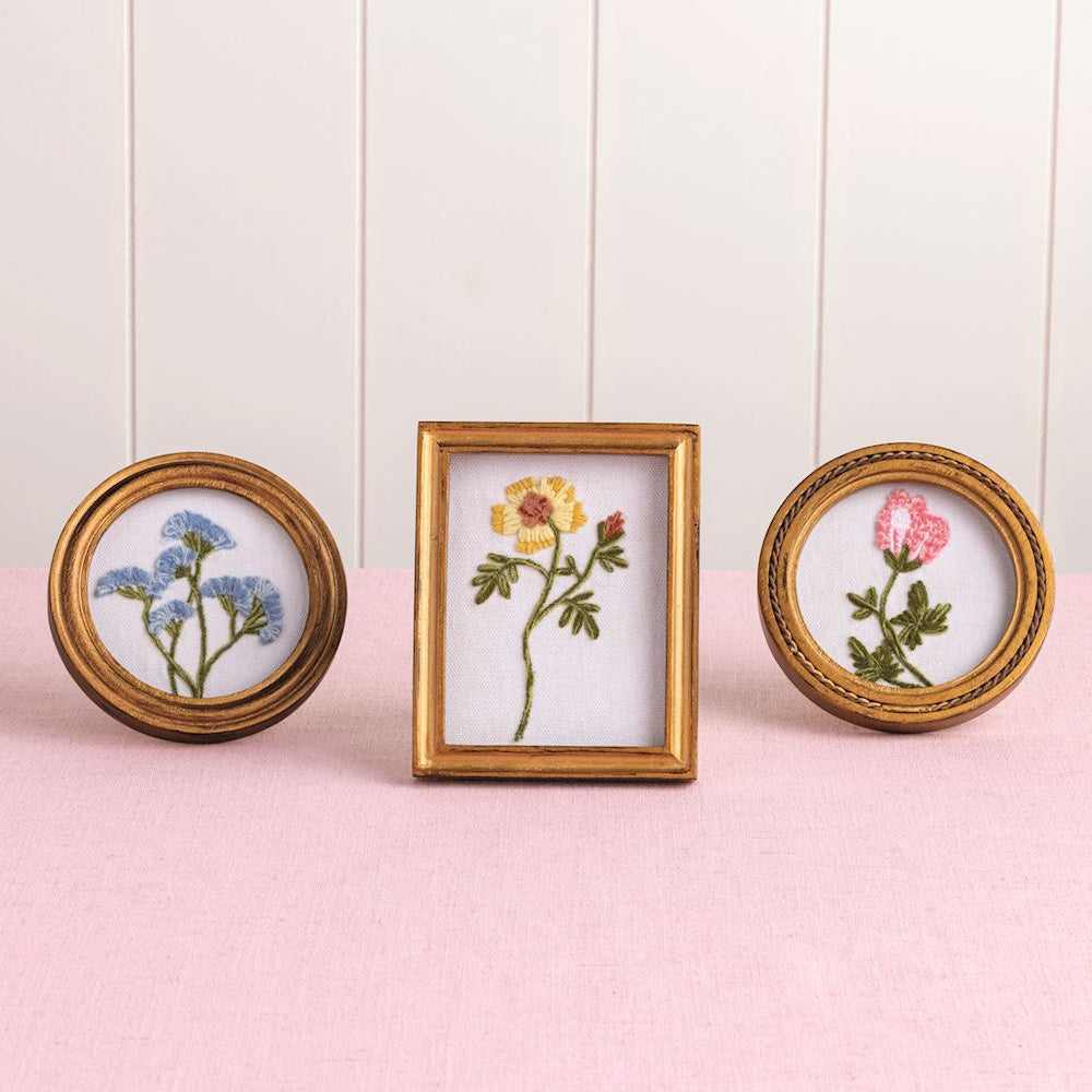 Three miniature gold-framed floral embroideries featuring blue, yellow, and pink wildflowers on a pink surface.
