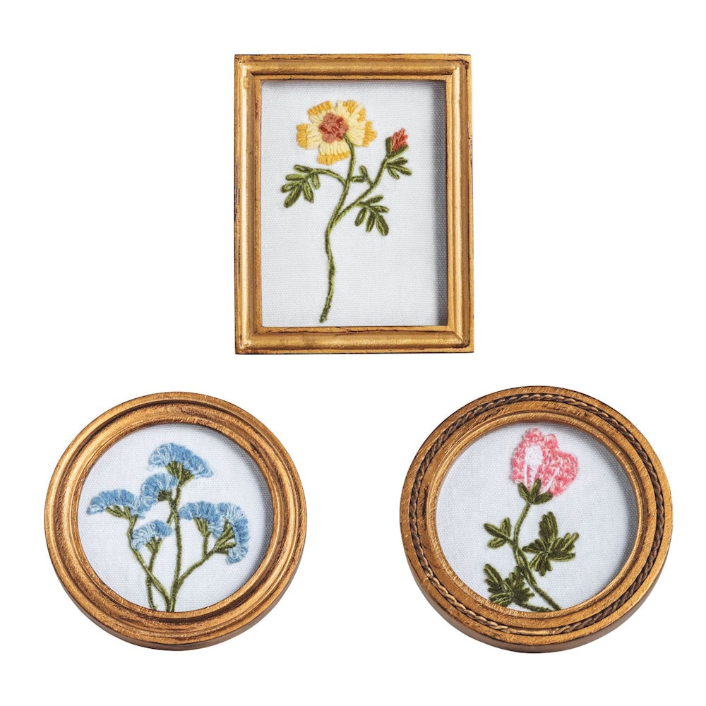 Three small gold-framed floral embroideries featuring yellow, blue, and pink wildflowers on a white fabric background.