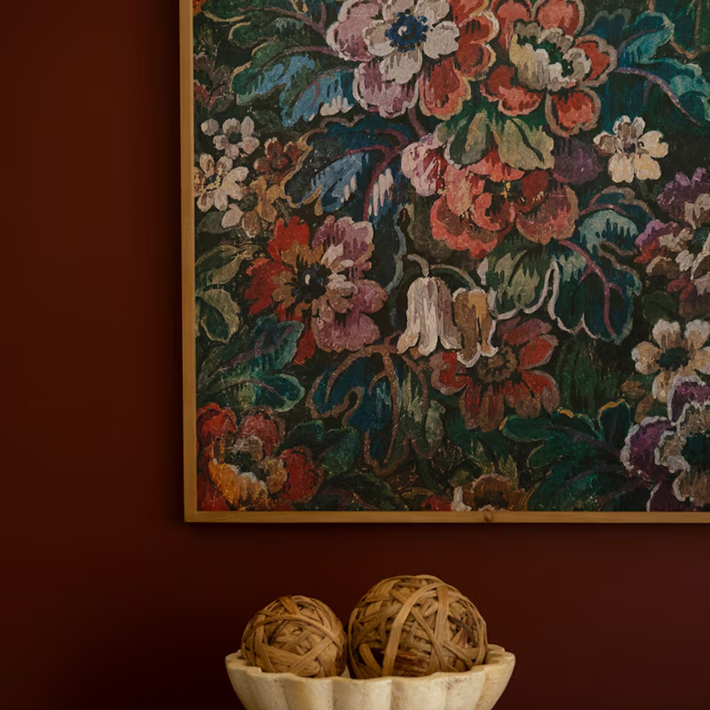 Vintage floral wall art in a gold frame above a bowl of decorative woven spheres on a dark moody red wall.
