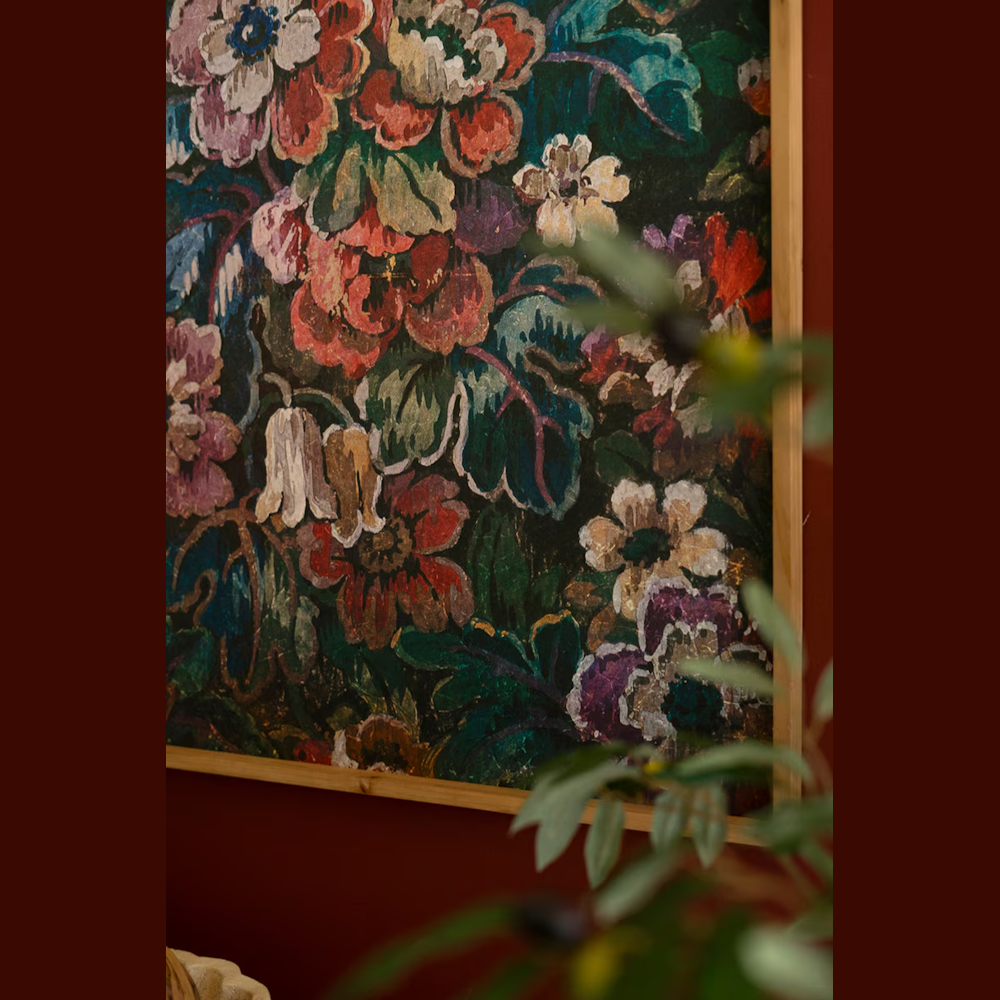 Vintage-style floral tapestry art in a light wood frame, featuring rich red and green botanical patterns on a dark wall.