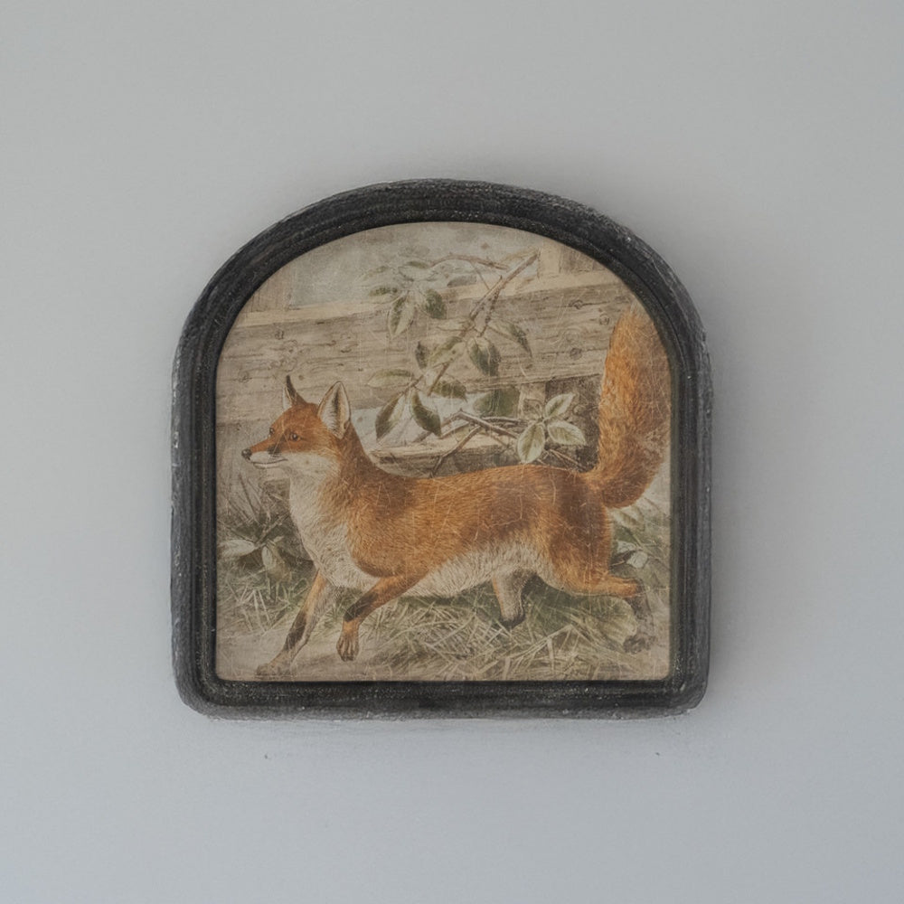 A vintage illustration of a red fox in an arched dark frame, set against a plain wall.