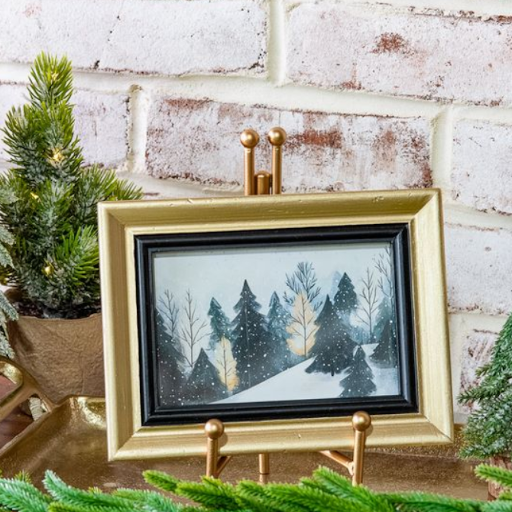 Framed painting of snowy pine trees displayed on a gold stand against a white brick wall.