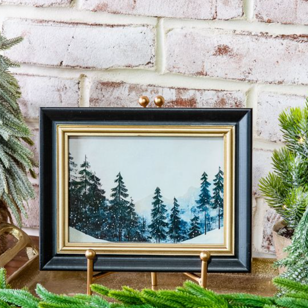 Framed winter landscape painting depicting pine trees on a gold stand against a white brick wall.