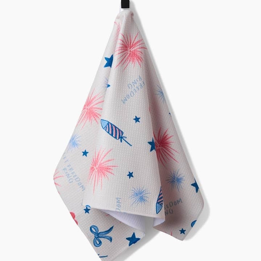 A white tea towel with red and blue illustrations of fireworks, stars, rockets, and "Let Freedom Ring" text.