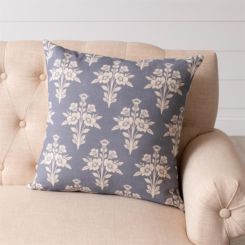 French blue square throw pillow with white floral medallion pattern on a beige tufted sofa.