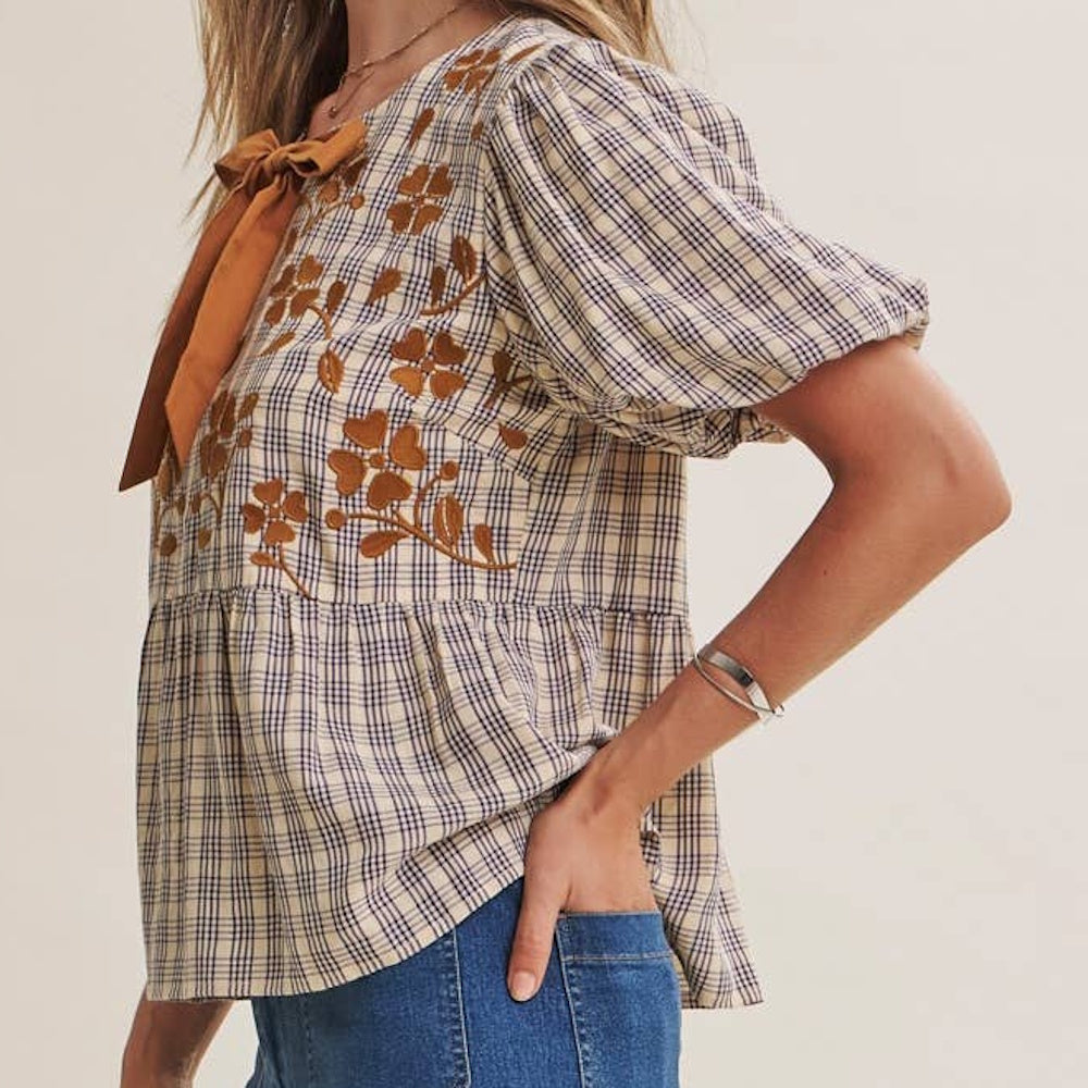 Side profile of plaid babydoll blouse with floral embroidery, short puff sleeves, and a orange neck tie.